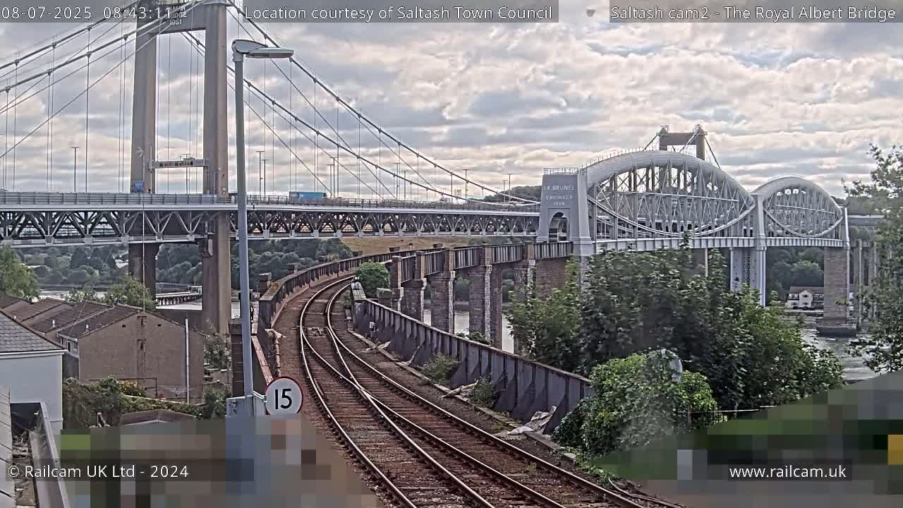 Train tracks curve toward a large suspension bridge and a smaller, arched bridge under partly cloudy skies.