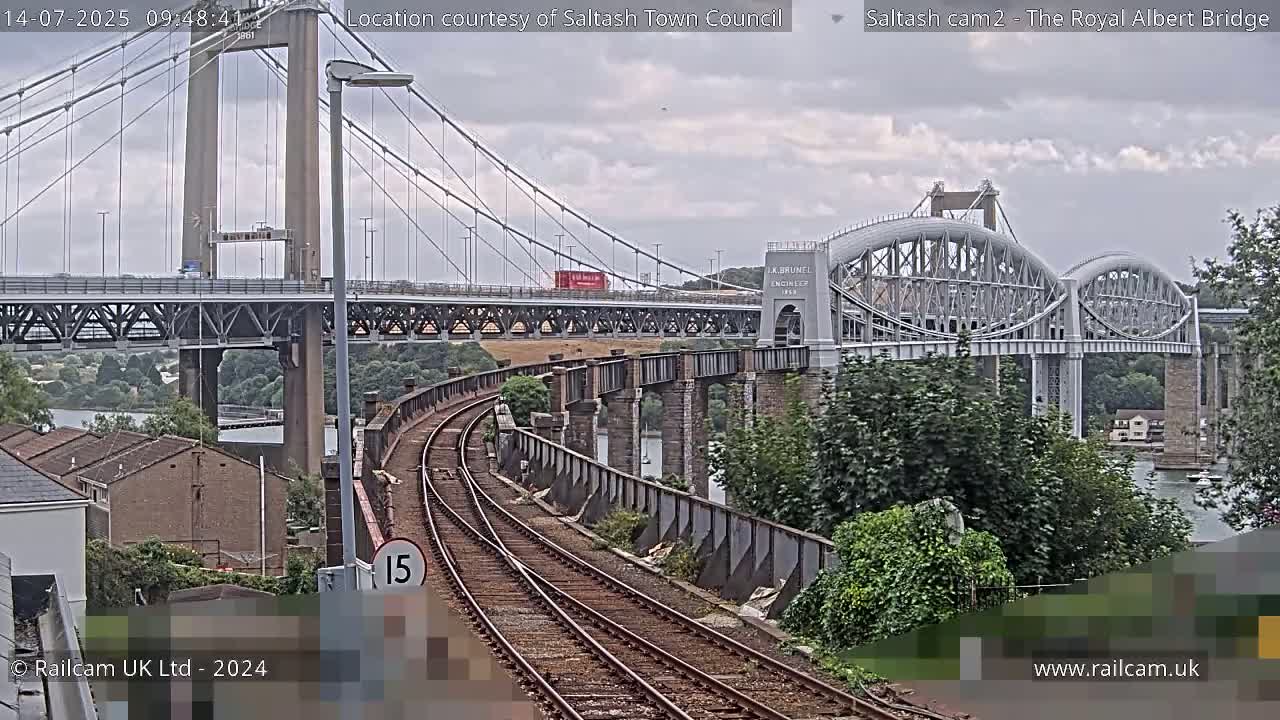 A view of a railway line curving towards two bridges, one a suspension bridge and the other an arch bridge, under an overcast sky.