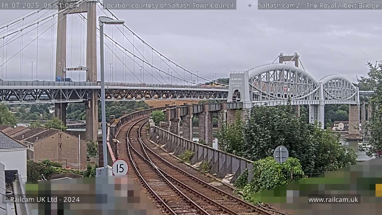 A train track curves towards two bridges, a suspension bridge and an arch bridge, under an overcast sky.