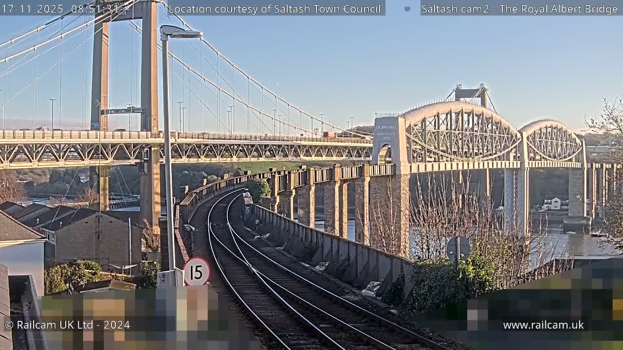 On a clear, sunny day, a curving railway line leads towards the confluence of a modern suspension bridge and an older arched railway bridge spanning a body of water.