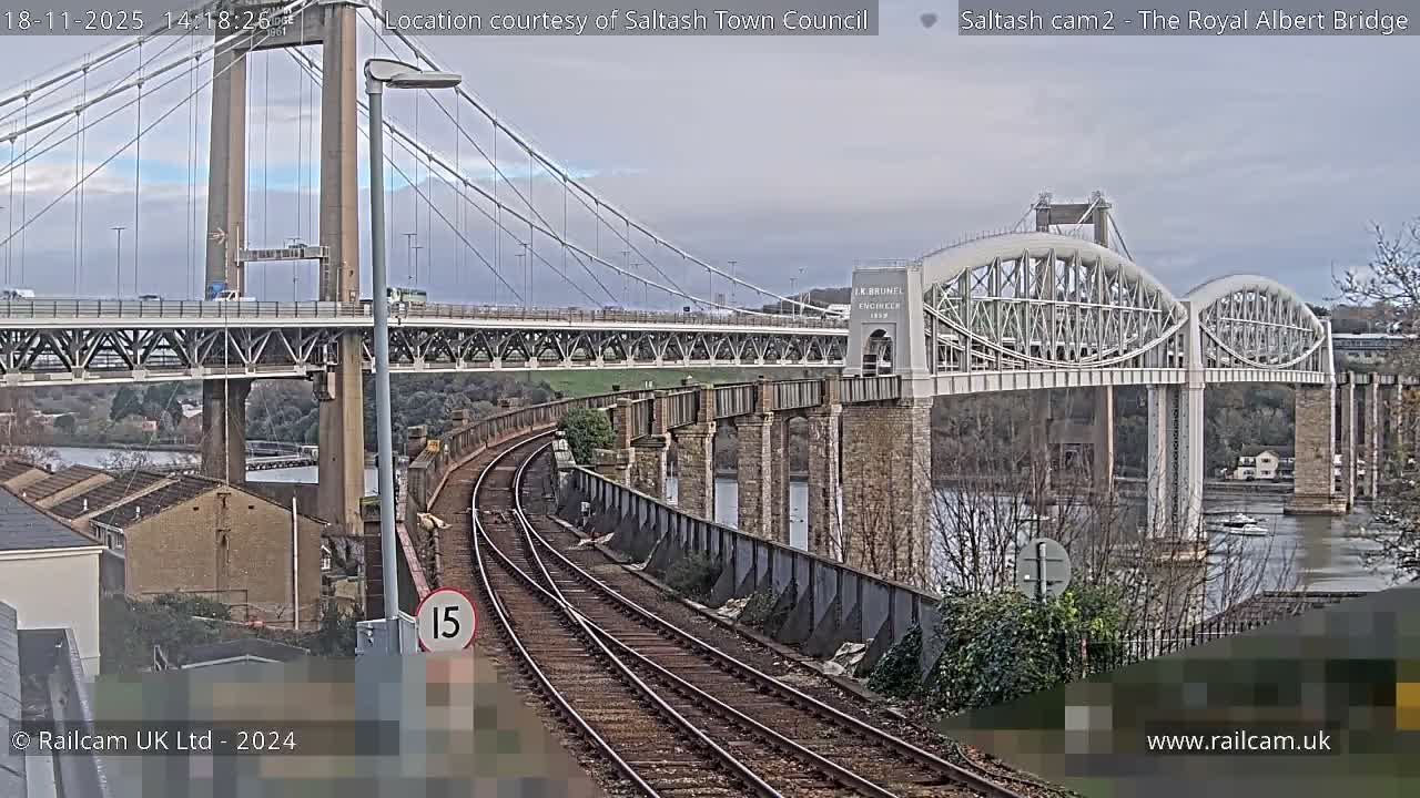 On a clear, sunny day, a curving railway line leads towards the confluence of a modern suspension bridge and an older arched railway bridge spanning a body of water.