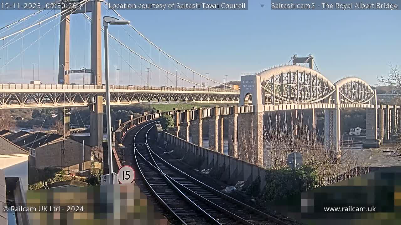 On a clear, sunny day, a prominent road suspension bridge and an arched railway bridge span a waterway, with curved train tracks and buildings in the foreground.