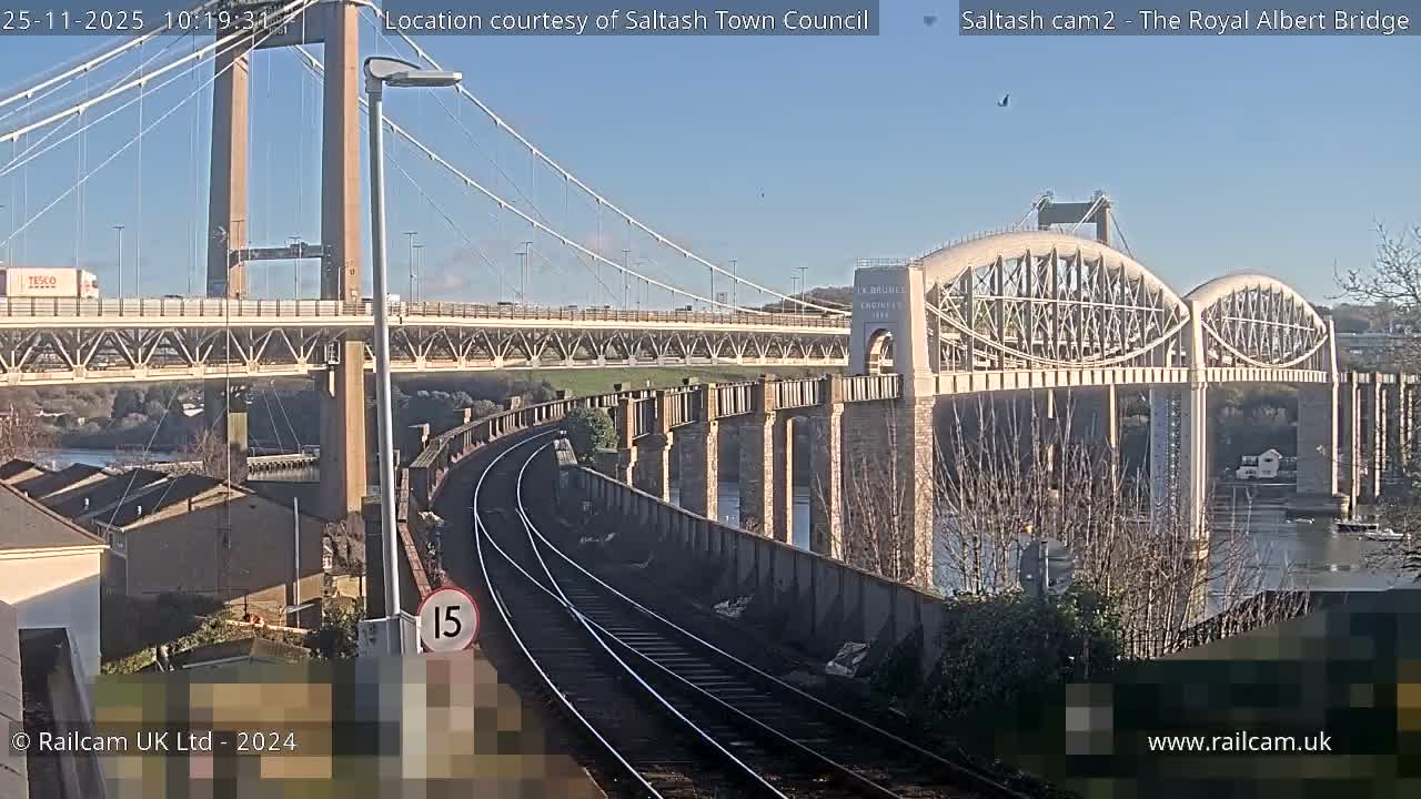 Under a clear blue sky, a sunny outdoor view reveals a complex of bridges, including the arched Royal Albert Bridge, spanning a body of water, with curving railway tracks prominent in the foreground.