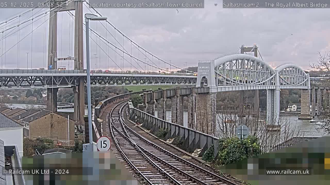 A wide view captures two prominent bridges, one a suspension bridge carrying road and rail and another arched railway bridge, spanning a river next to curving railway tracks and various buildings under an overcast sky.