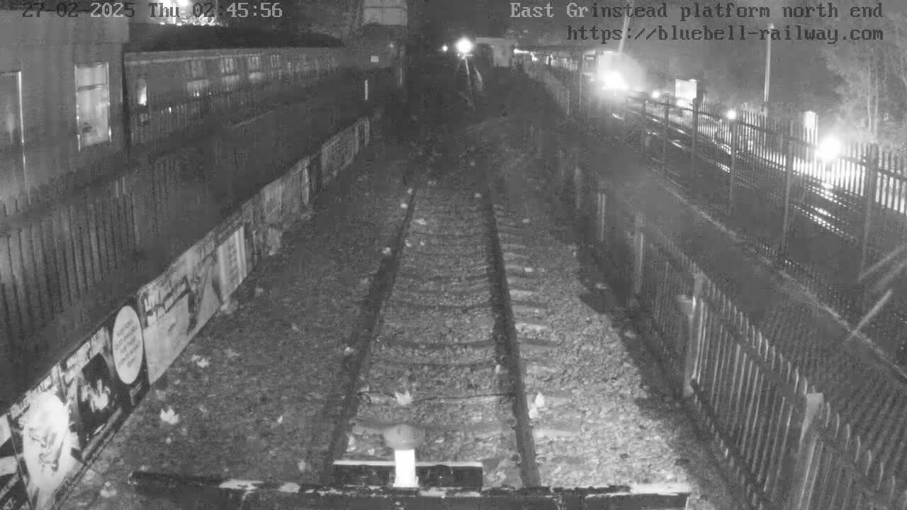 A nighttime view of train tracks at a station platform, with a train car visible to the left and fences on either side.