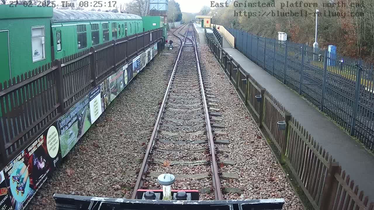 A view from a train shows a railway track extending to a station platform, bordered by fences and posters, on an overcast day.