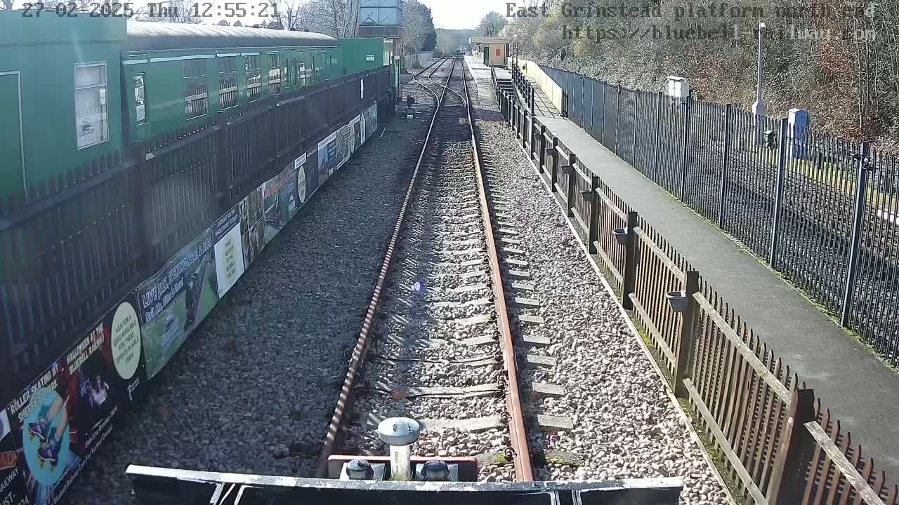 A view from a train's perspective shows train tracks extending into the distance on a sunny day, alongside a platform with a green train car and fences.
