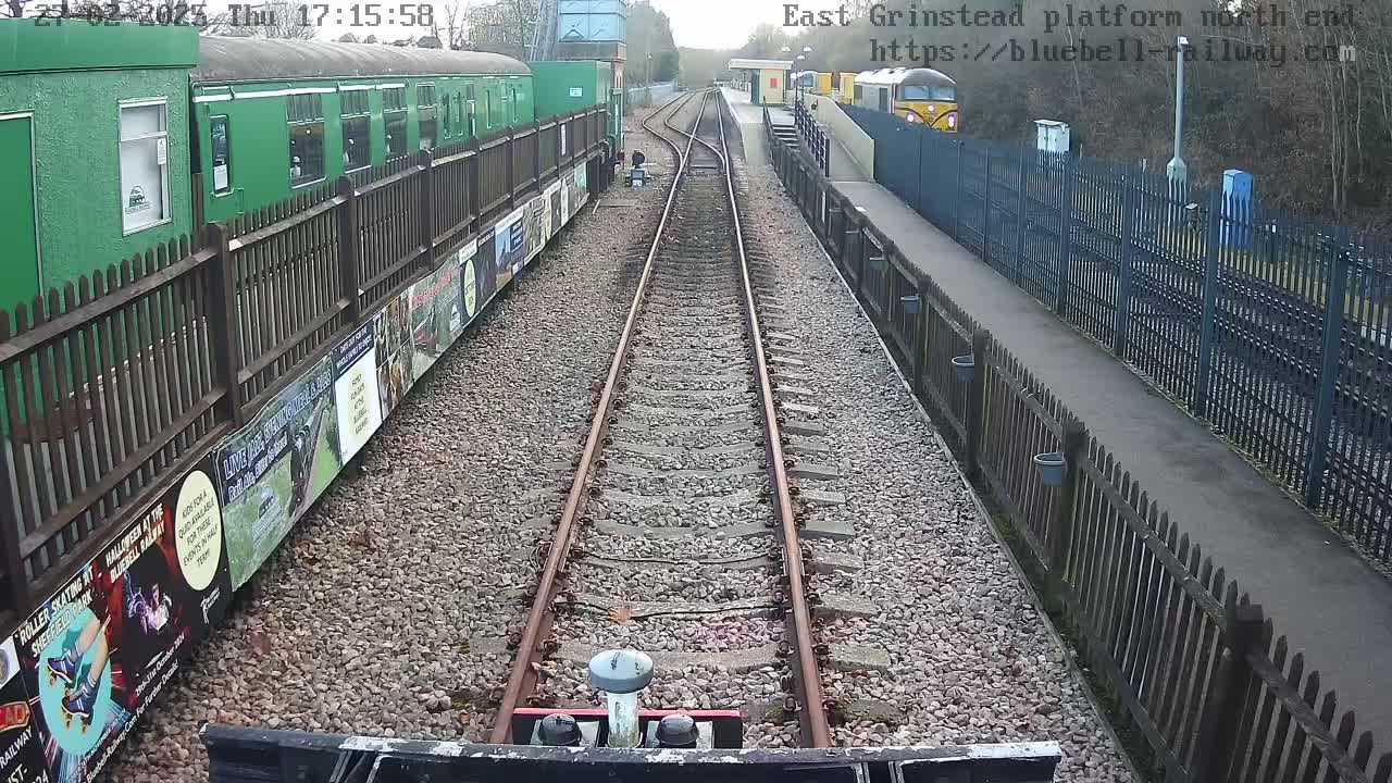 A view from a train along a railway track, with a green train car on the left and a platform and another train visible in the distance, on a partly cloudy day.