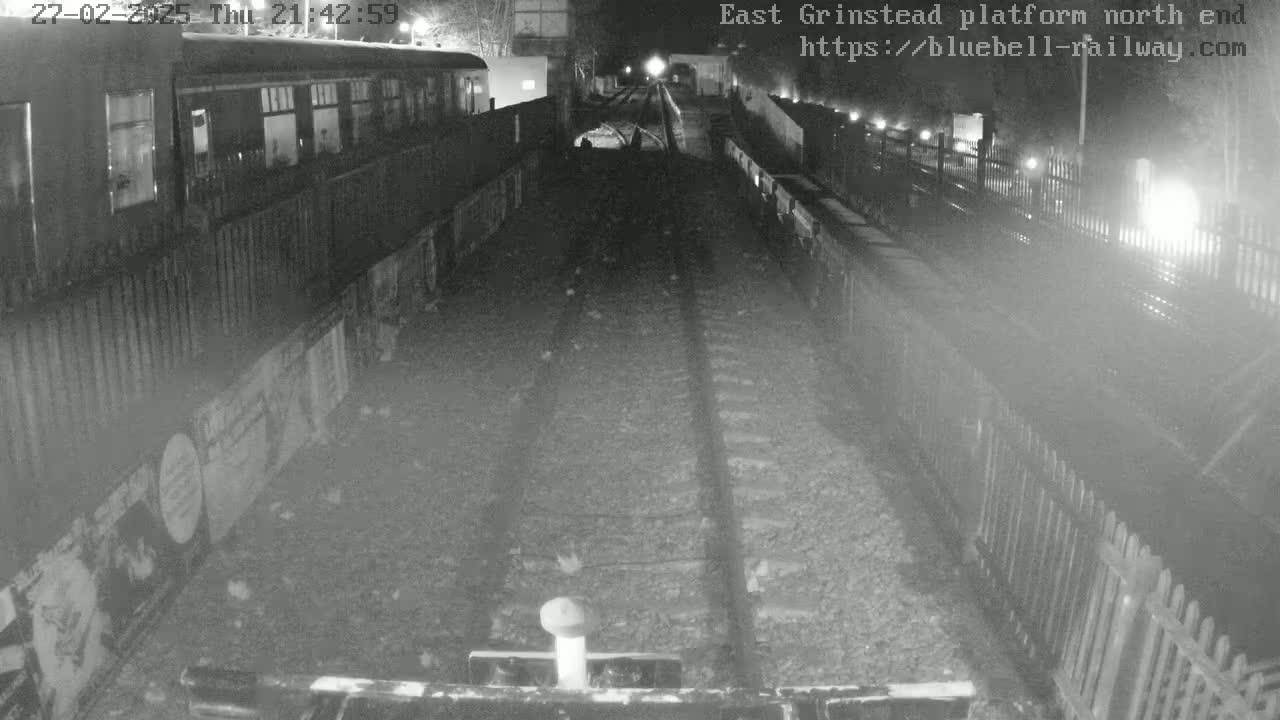 A nighttime view of train tracks at a station platform, with a train partially visible and a tunnel in the distance.