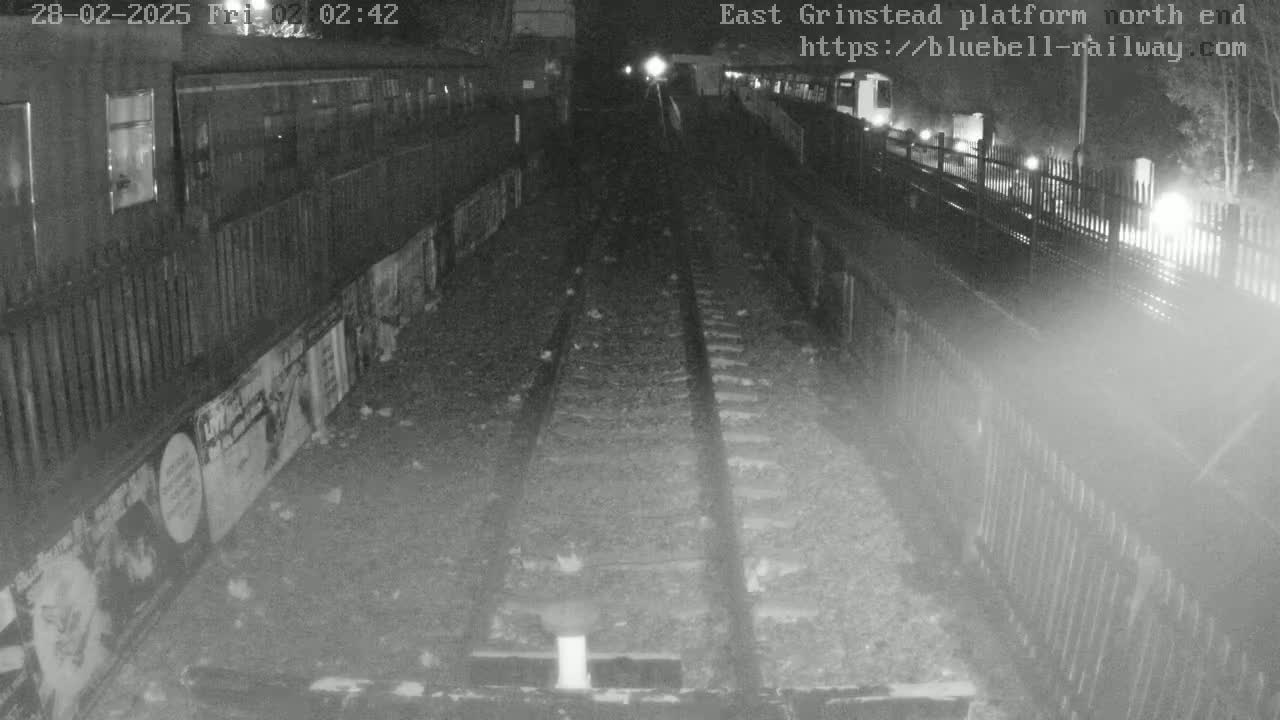 A nighttime view of train tracks at a platform, with a train car partially visible and posters lining a wall beside the tracks.