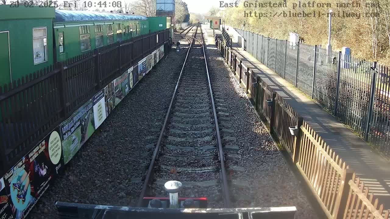 A view from a train's perspective shows double railway tracks stretching to a platform bordered by fences and posters, under a sunny sky.