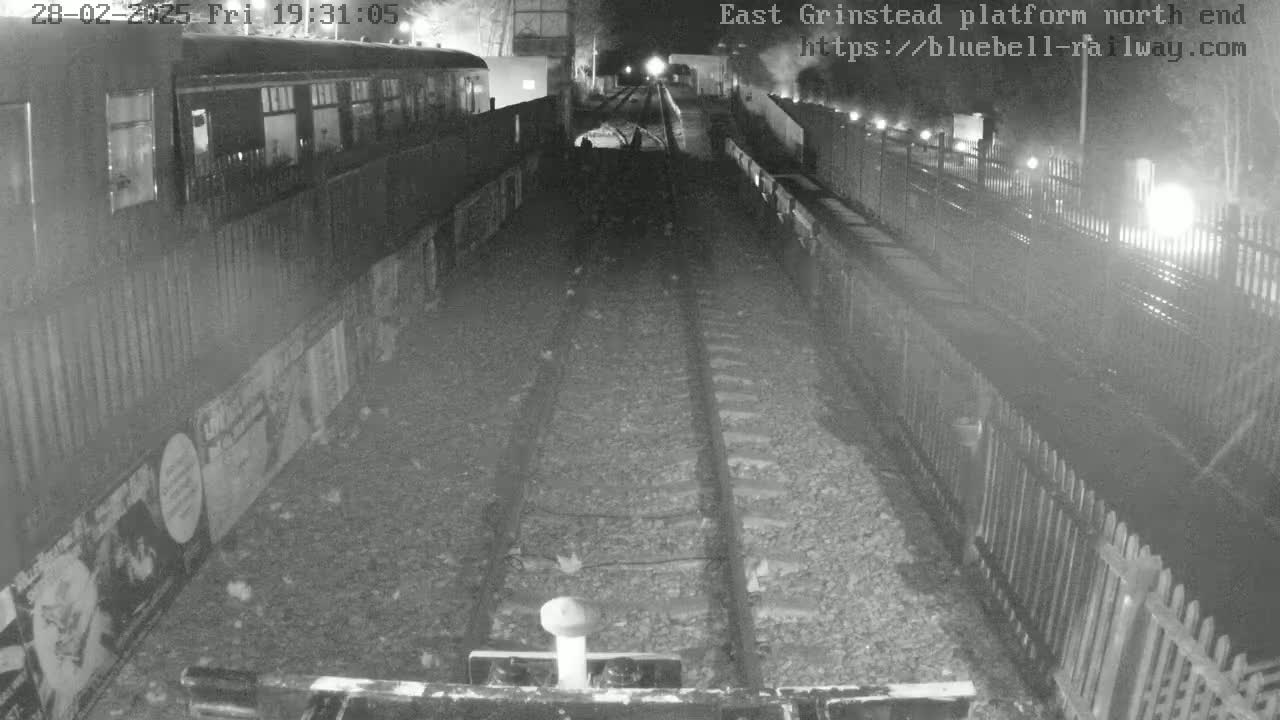 A nighttime, low-light view of train tracks extending into the distance between platforms, with a train car visible on the left and fencing along the sides.