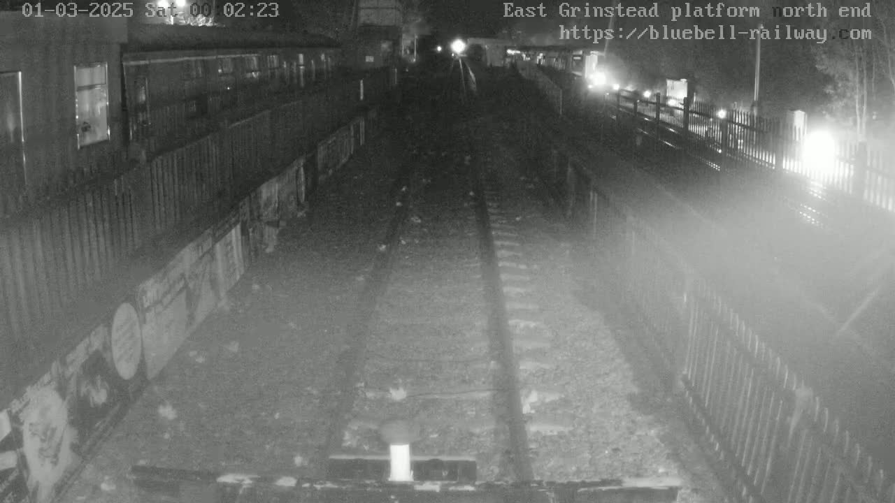 A nighttime view of train tracks at a dimly lit railway platform, with a train partially visible in the background.