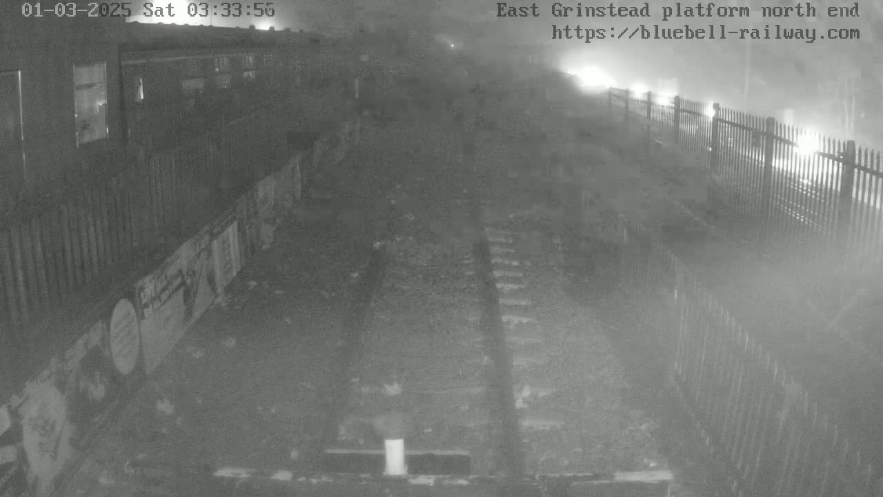 A nighttime, foggy view of a train car partially visible next to railway tracks bordered by a fence.