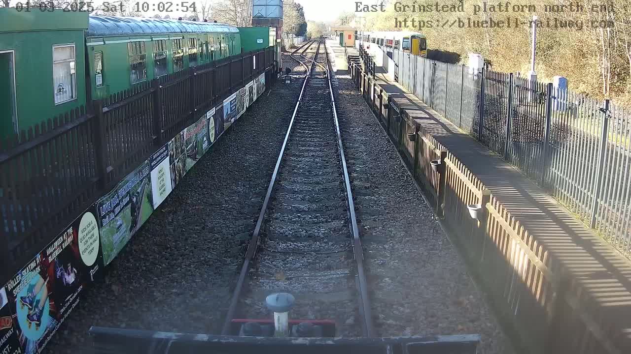 A sunny view down a railway track, between two platforms with green train cars and fences lined with posters.