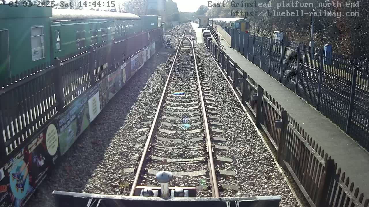 A sunny day view from a train's perspective, showing railway tracks extending toward a station platform bordered by fences and a green train car.