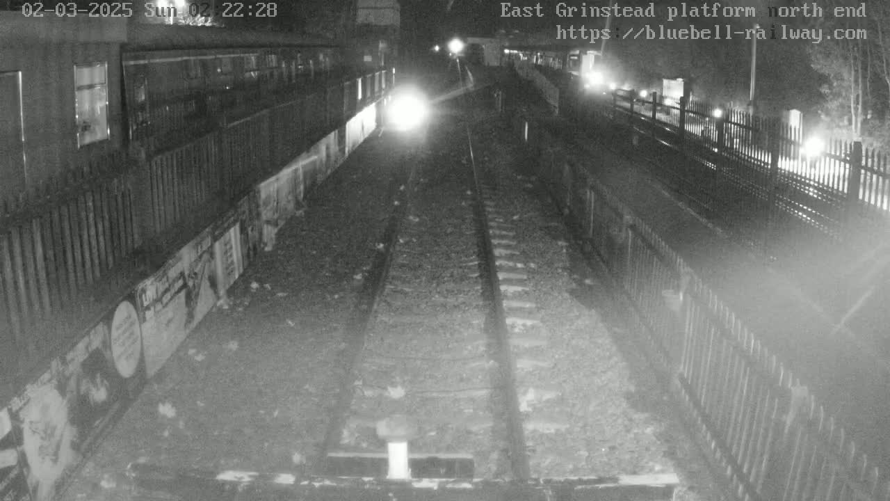 A nighttime view of train tracks at a platform, with fences and posters lining the sides.