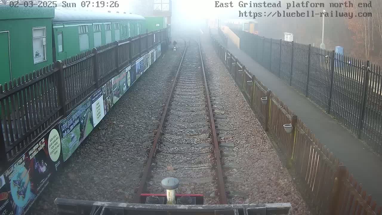 A foggy view down a railway track, bordered by fences and advertisements, with a green train car visible to the left.