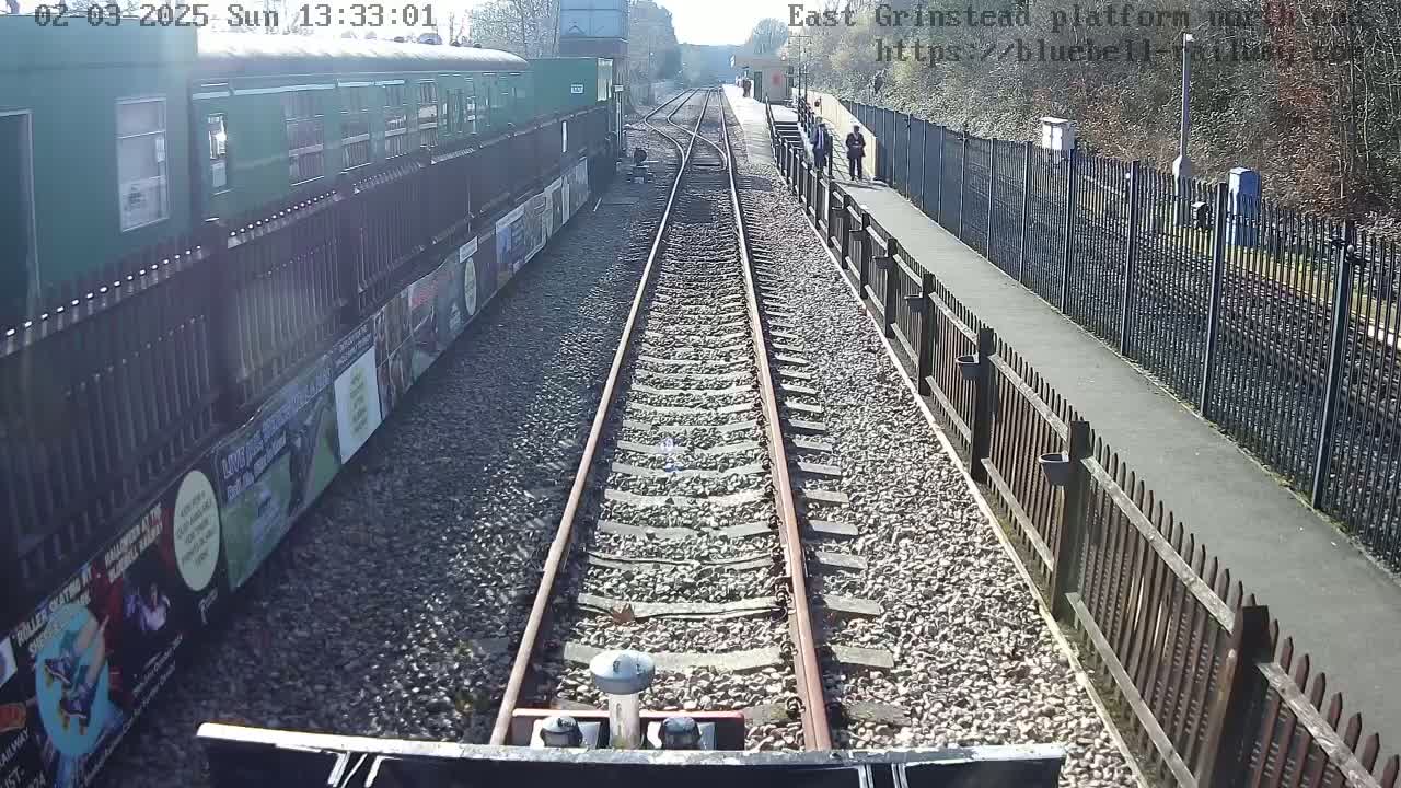 A view from a train on a sunny day shows two parallel train tracks running alongside a platform with a fence, a green train car, and a paved walkway.