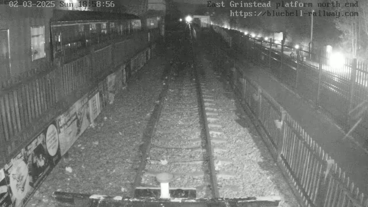 A nighttime, low-light view of a train platform shows railroad tracks extending into the distance between fences, with posters lining one side of the platform.