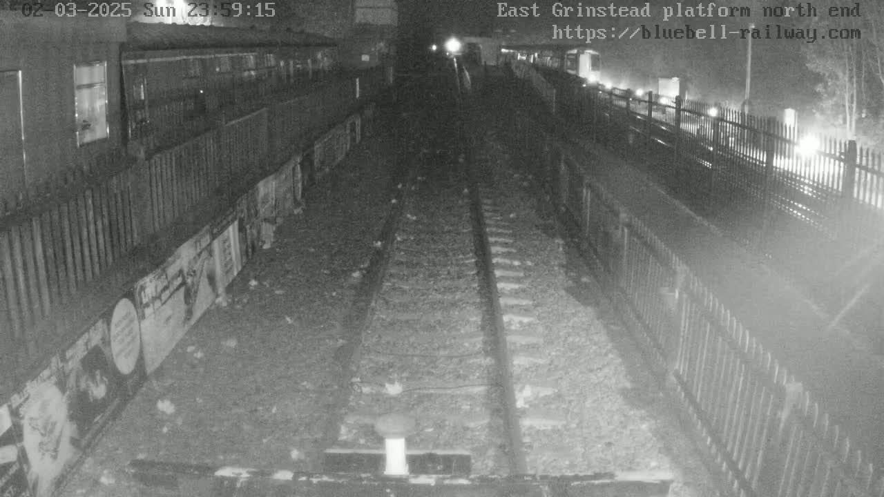 A nighttime view of a train platform's north end, showing empty train tracks and a fence along the platform edge.