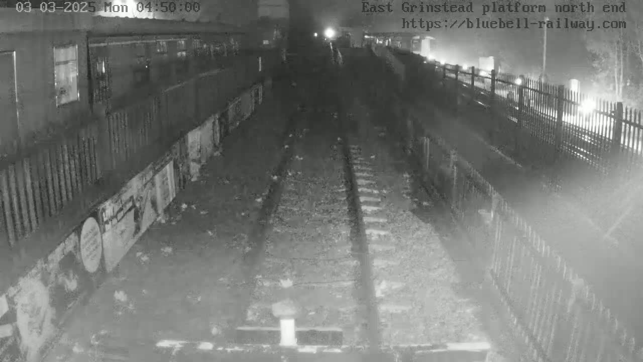 A nighttime, low-light view of train tracks extending into the distance, bordered by a fence and a building with graffiti.