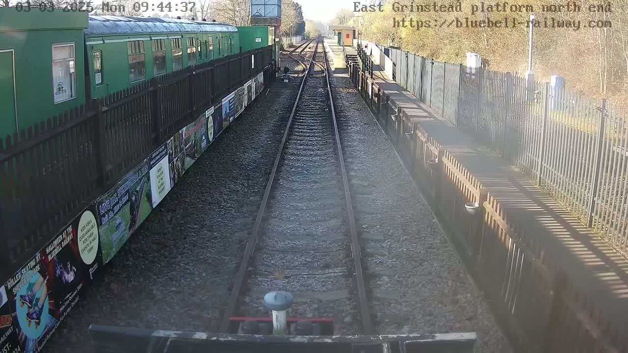 A view from a train on a sunny day shows a railway track extending to a station platform with a green train car and fences along the sides.