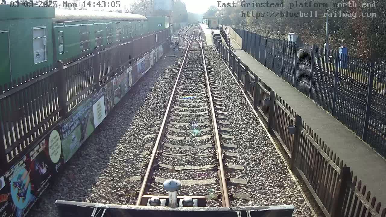 A sunny view from a train shows a railway track extending to a station platform bordered by fences and advertisements.