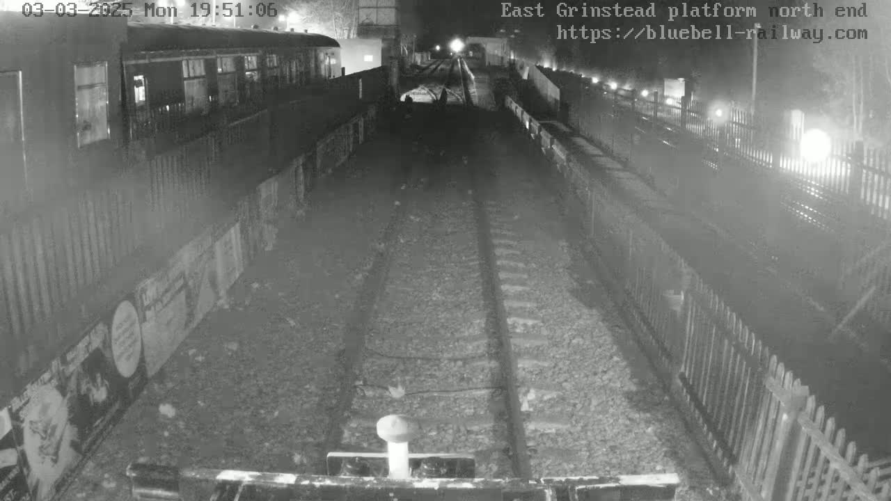 A nighttime view of train tracks extending into the distance between low fences, with a train car partially visible on the left.