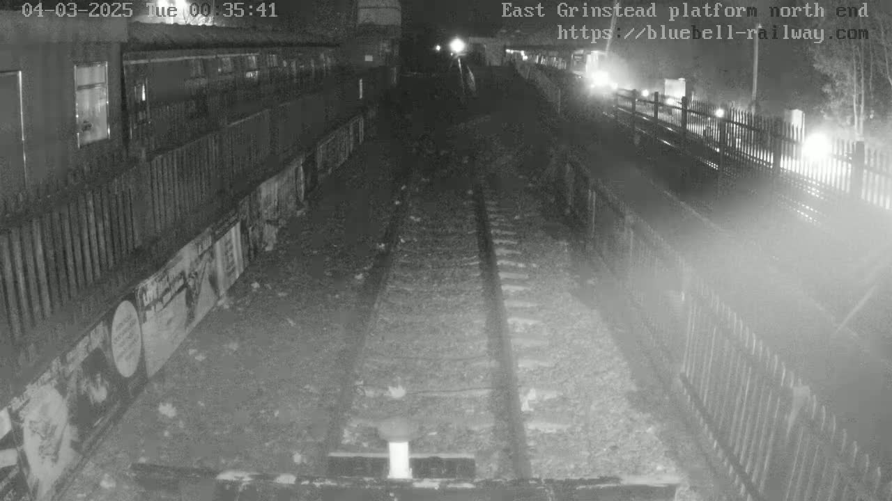 A nighttime view of train tracks at a station platform, bordered by fences and featuring posters on the wall.