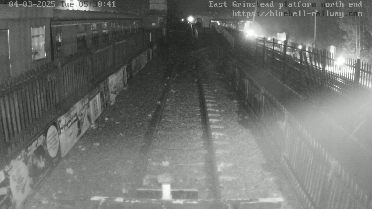 A nighttime, low-light view of train tracks at a platform, with fences and walls on either side.