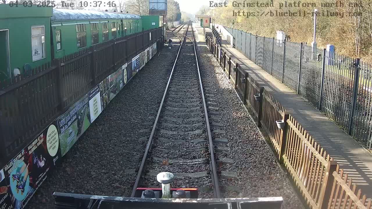 A view down a railway track on a sunny day, with a green train car and posters on the left and a wooden fence on the right.