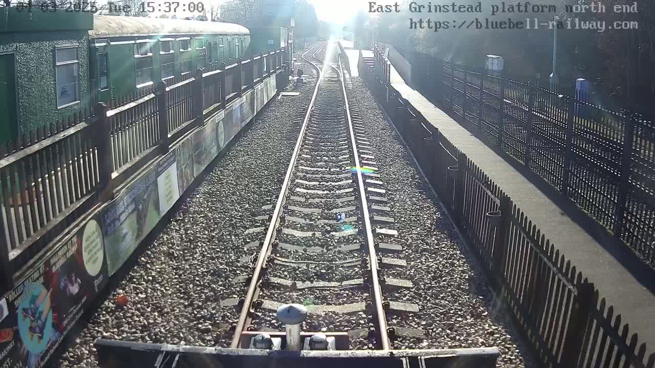 A sunny view down a railway track, alongside a platform with a green train car and fences.