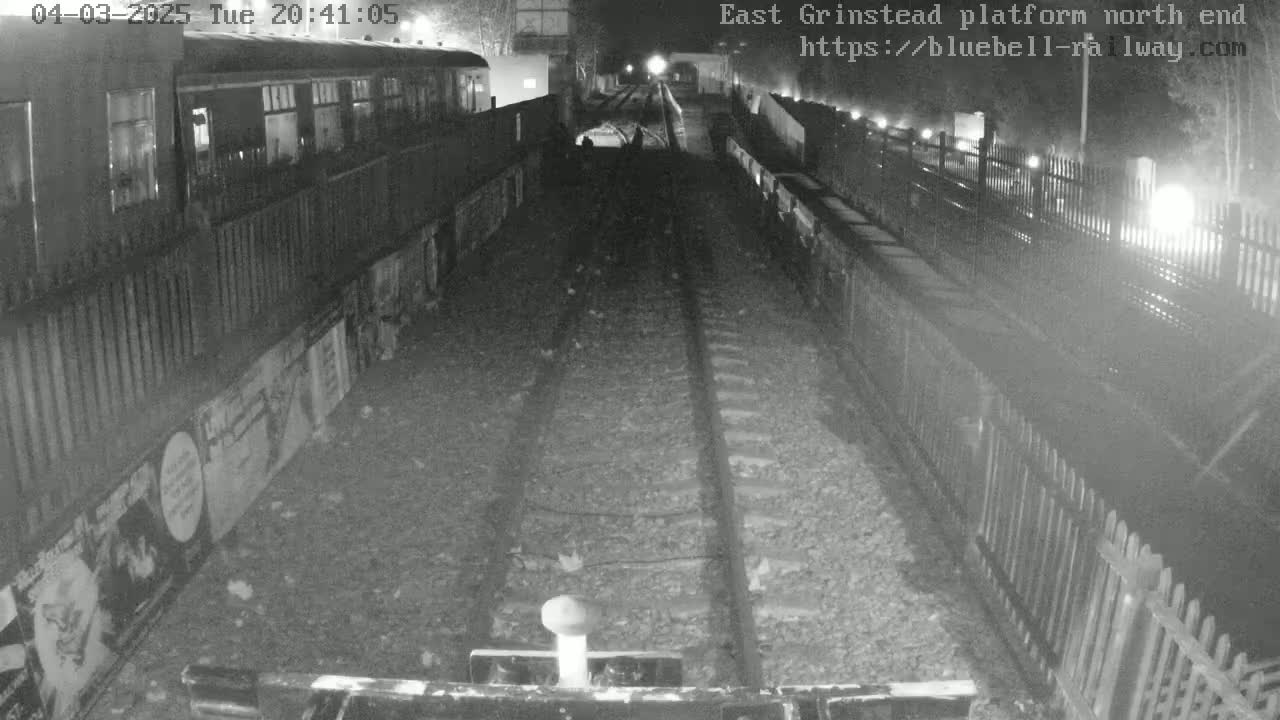 A nighttime view of train tracks at a station platform, with a train partially visible and fences lining the sides.