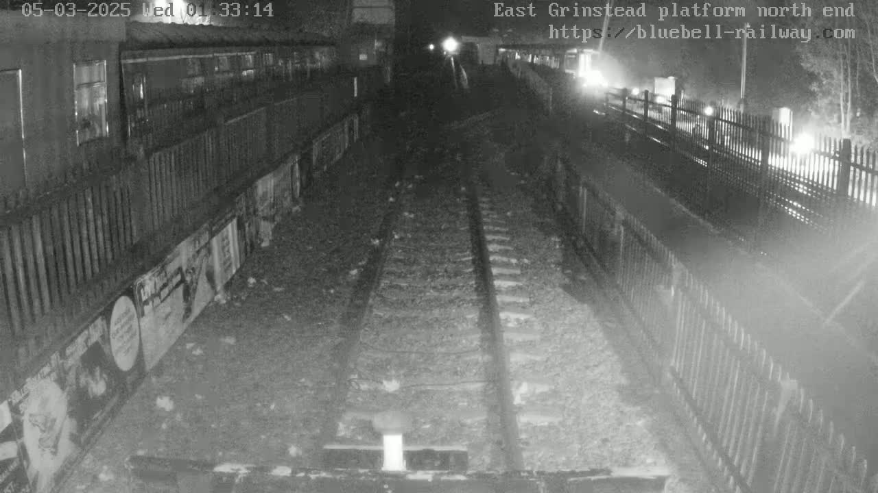 A nighttime view of train tracks at a platform, bordered by fences and lined with posters, under dark, possibly cloudy conditions.