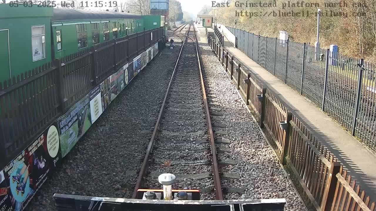 A view down a railway track on a sunny day, showing a green train car and wooden fences alongside the tracks.