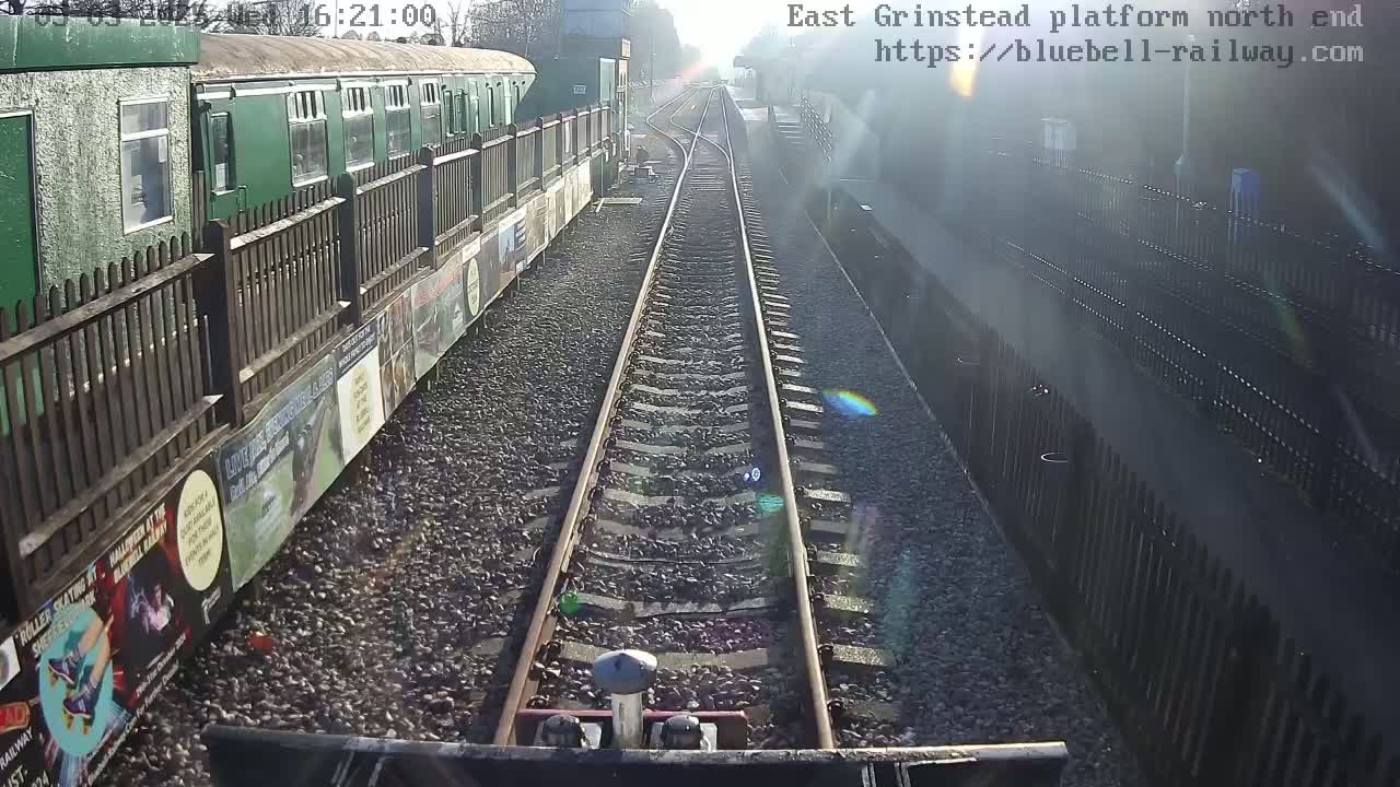A view from a train shows a sunny day at a railway platform, with a green train car and a wooden fence alongside the tracks.
