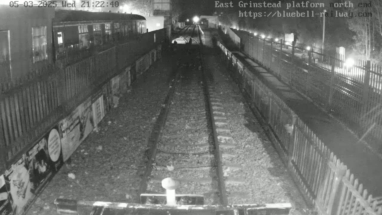 A nighttime, low-light view of train tracks extending into a dark tunnel, flanked by fences and a train car partially visible on the left.