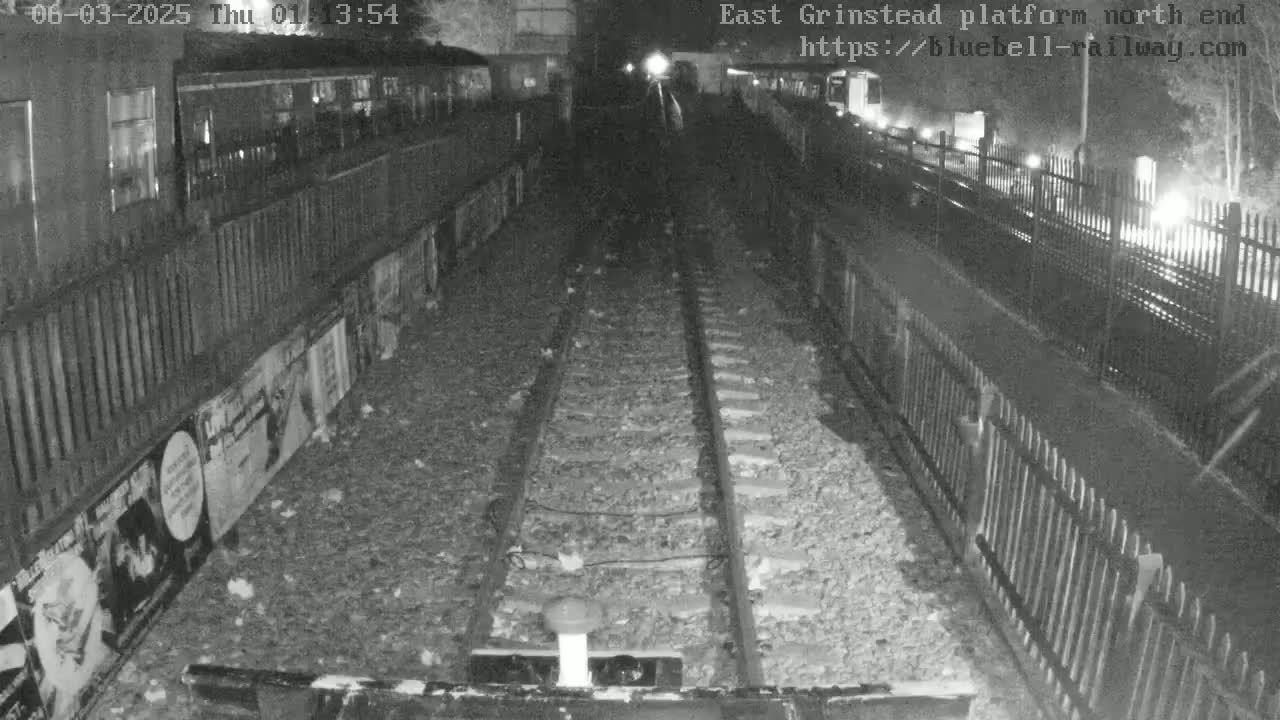 A nighttime, black-and-white view of train tracks extending into a tunnel, bordered by fences and posters.