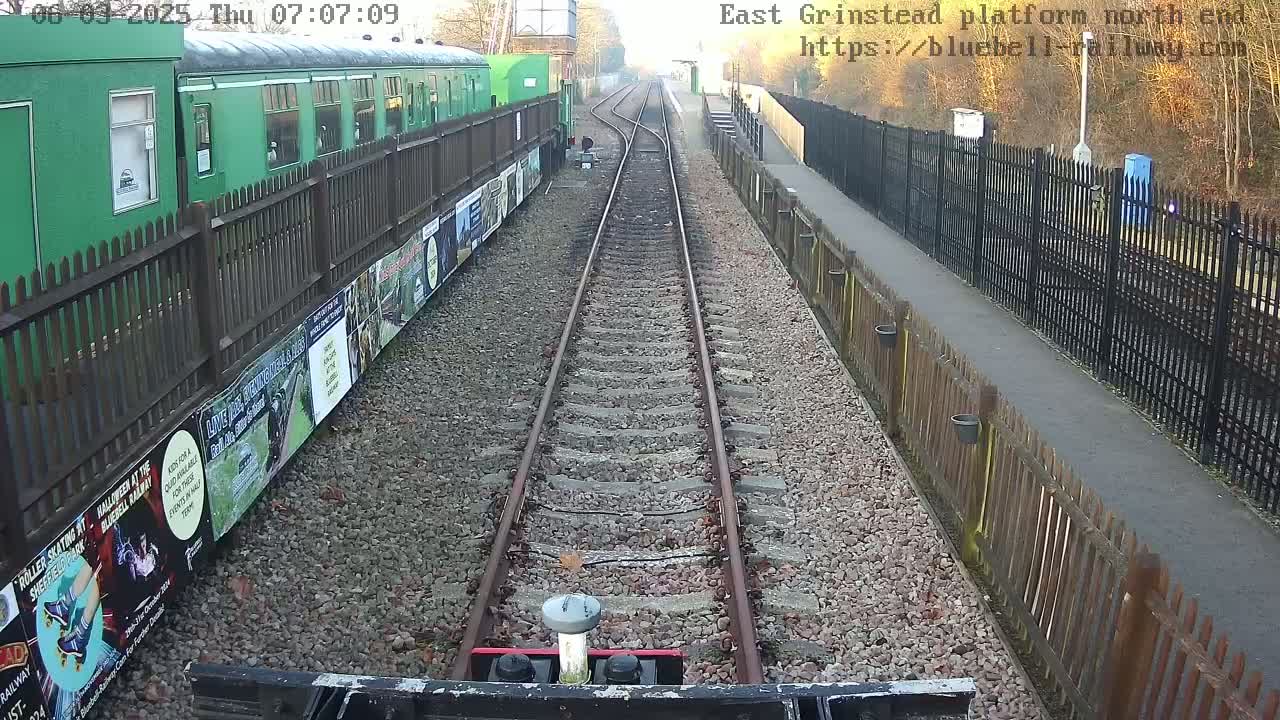 A view from a train shows a railway track extending to a station platform with green rail cars and fences on either side, under a partly sunny sky.
