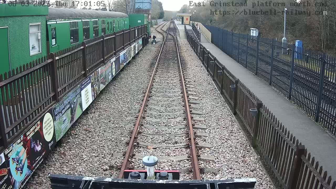 A view from a train on a railway track, showing a green train car and a platform with fences on a mostly clear day.