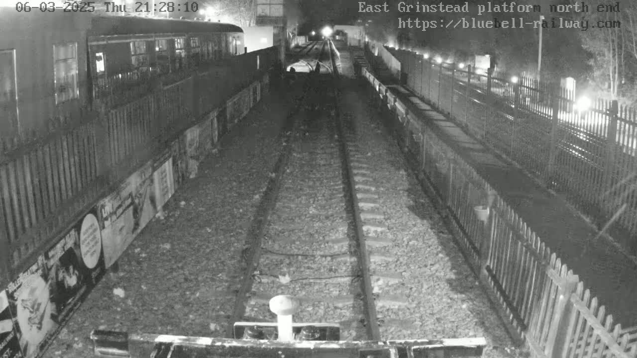 A nighttime, low-light view of train tracks at a station platform with a train car visible to the left and fences lining the sides.