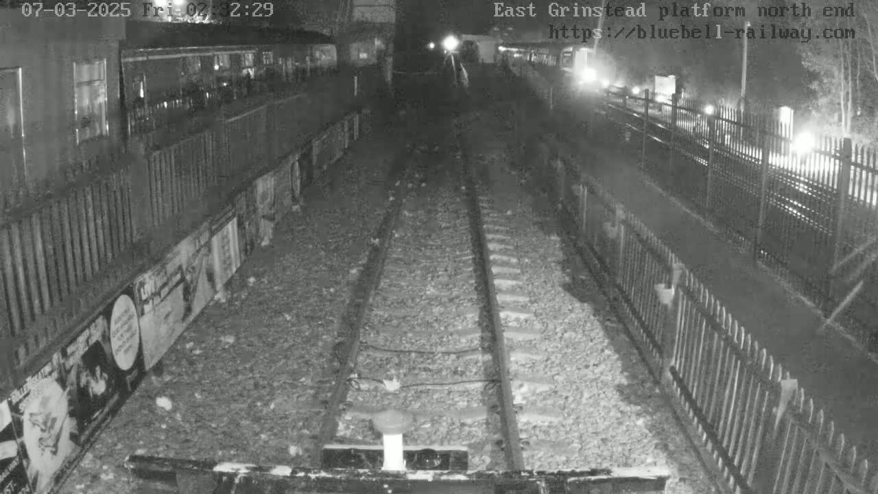 A nighttime view of a train platform's north end, showing train tracks and a fence, with low-light conditions.