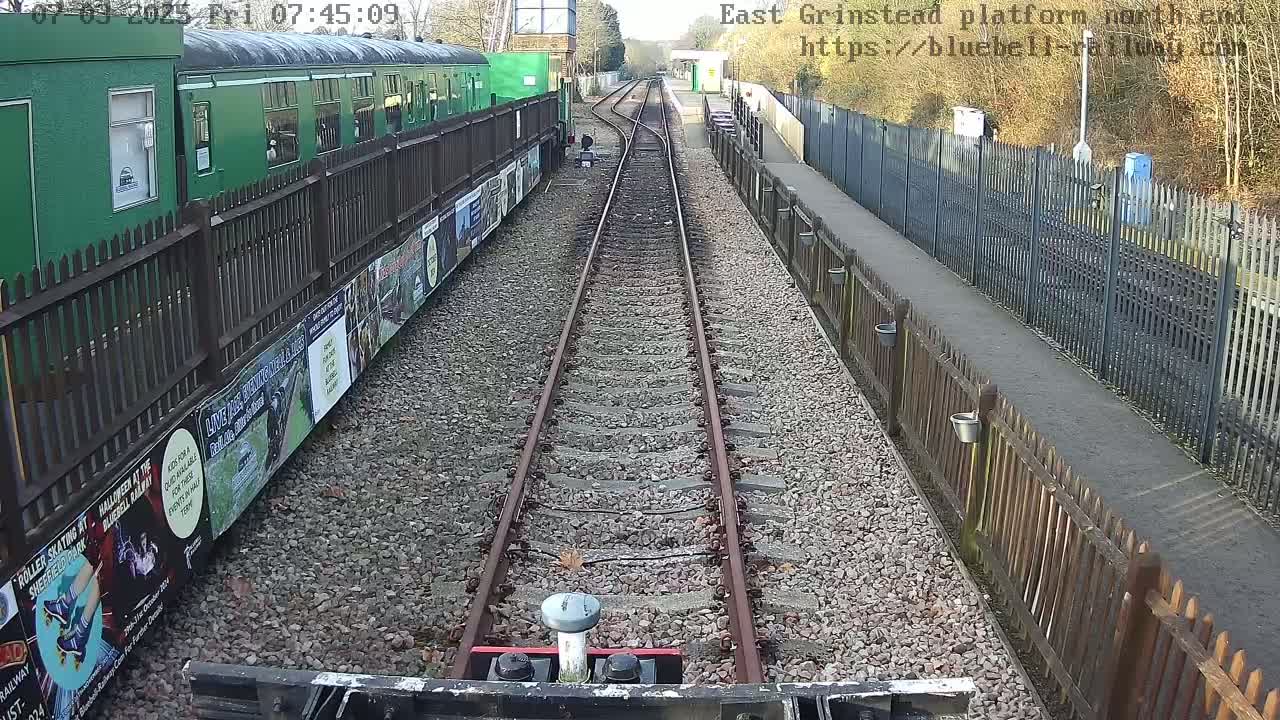 A view down a railway track on a sunny day, with a green train car visible to the left and wooden fences along both sides of the track.