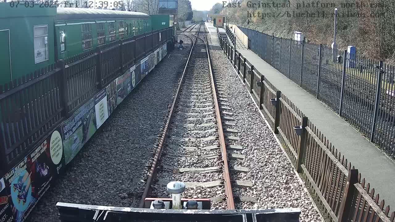 A view from a train on a sunny day shows railway tracks extending toward a station with green railcars alongside a wooden fence and posters on the side.