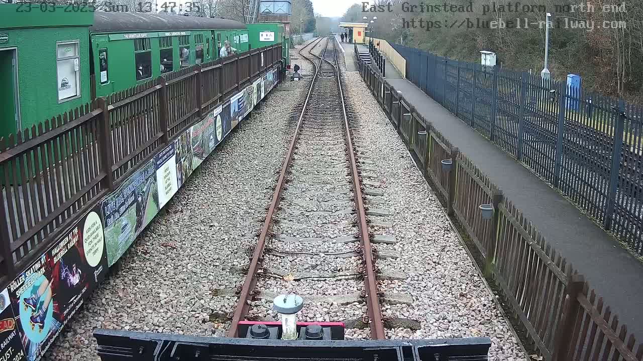 A view from a train's perspective shows a railway track extending to a platform with a green train car and wooden fences on either side, under a cloudy sky.
