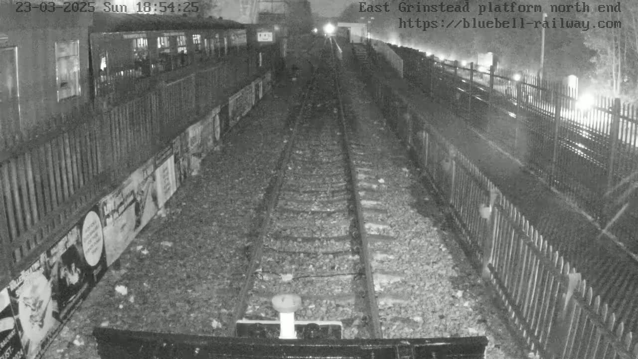 A nighttime view of a train platform's north end, showing train tracks stretching into the distance, alongside fences and posters on the walls.