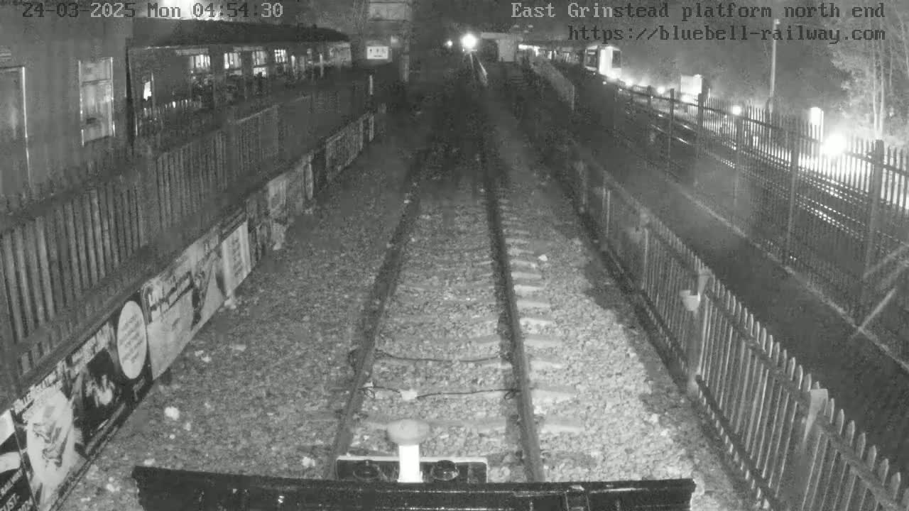 A nighttime view of a train platform shows two sets of train tracks extending into the distance between fences and posters on the walls.