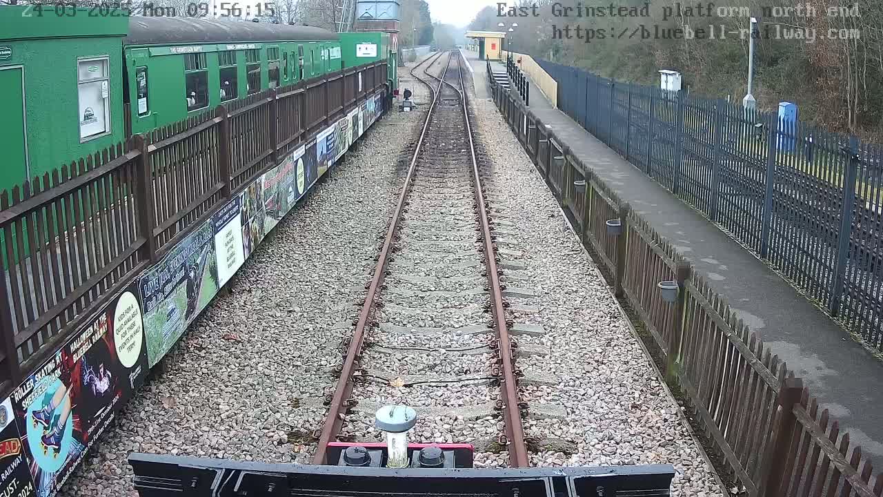 A view from a train's perspective shows a railway track extending into the distance, flanked by gravel ballast and wooden fences, with a green train car and posters visible to the left.