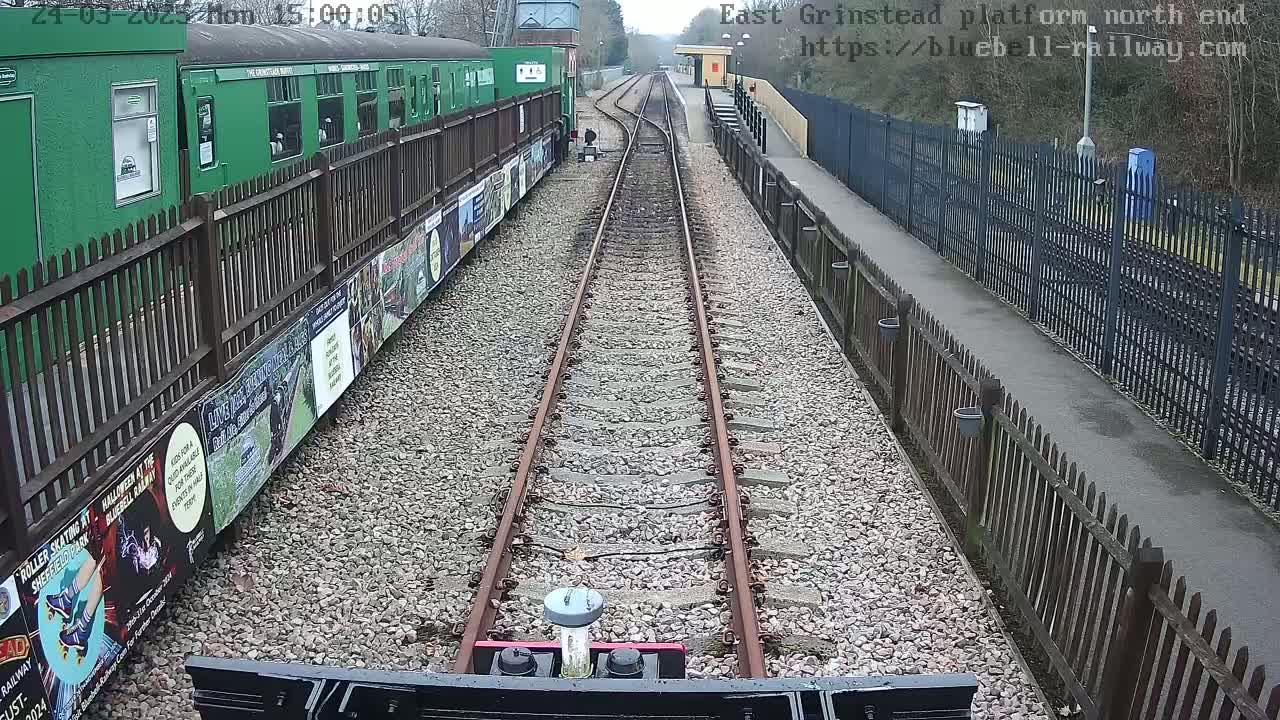 A view from a train's perspective down a railway track on a partly cloudy day, alongside a platform with a green train and wooden fences.